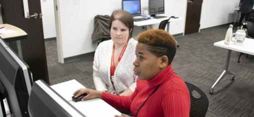 A woman in a red sweater looks at a computer while another woman helps her.