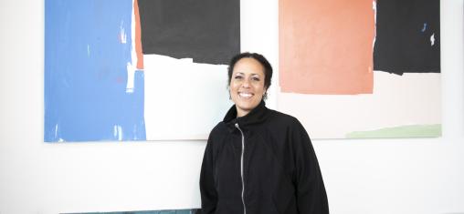 A woman, Akwi Nji, stands in front of her abstract paintings featuring blocks of color.