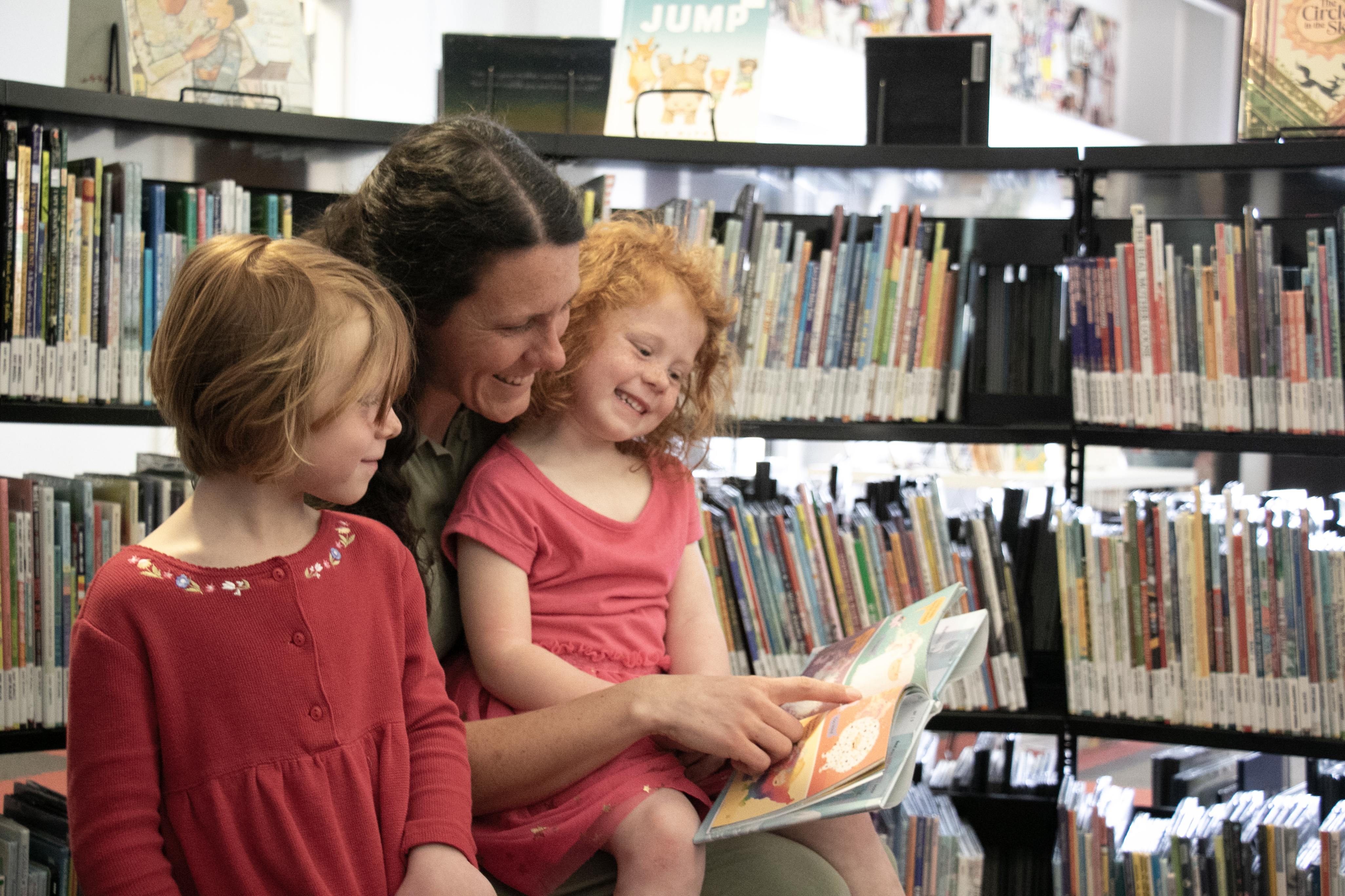 A woman reads to a girl on her lap while another girl looks on.