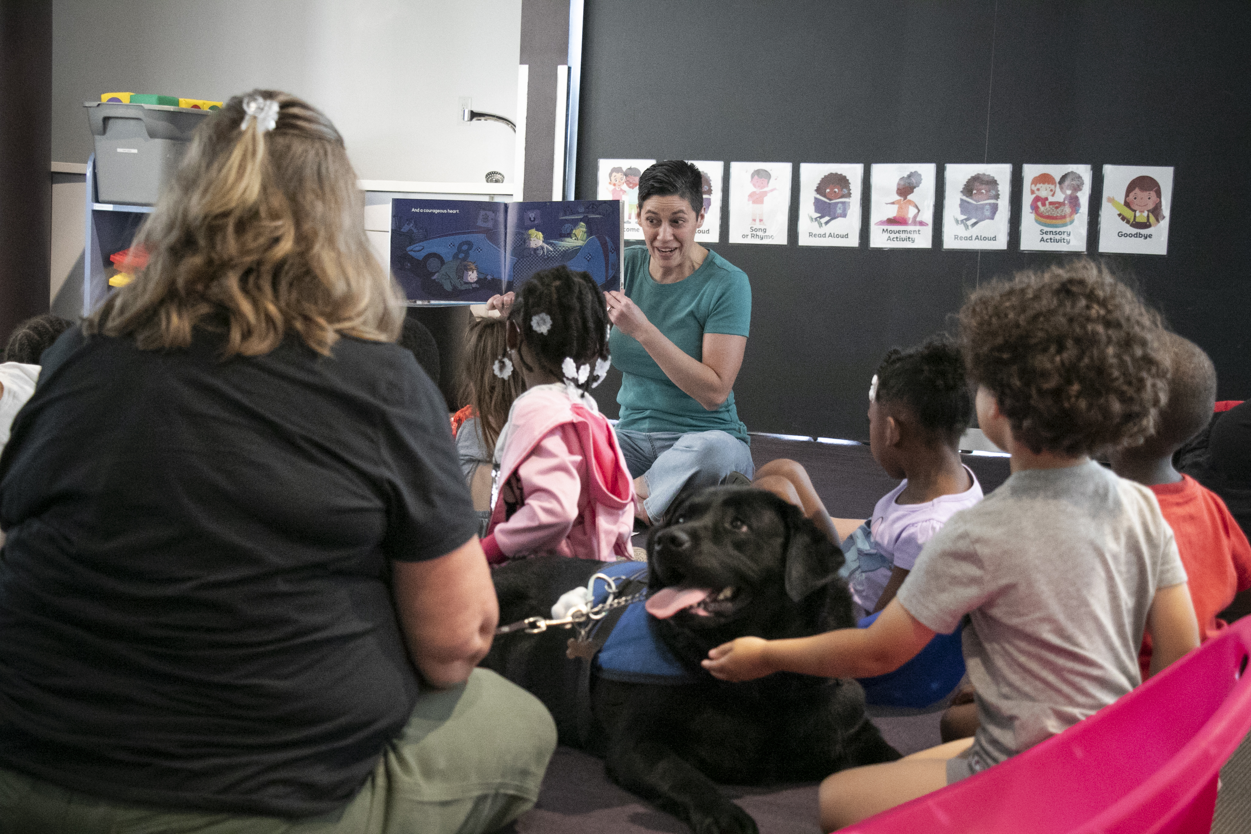 A woman reads a book to a room full of children and one dog, who wears a vest indicating he is a service dog.