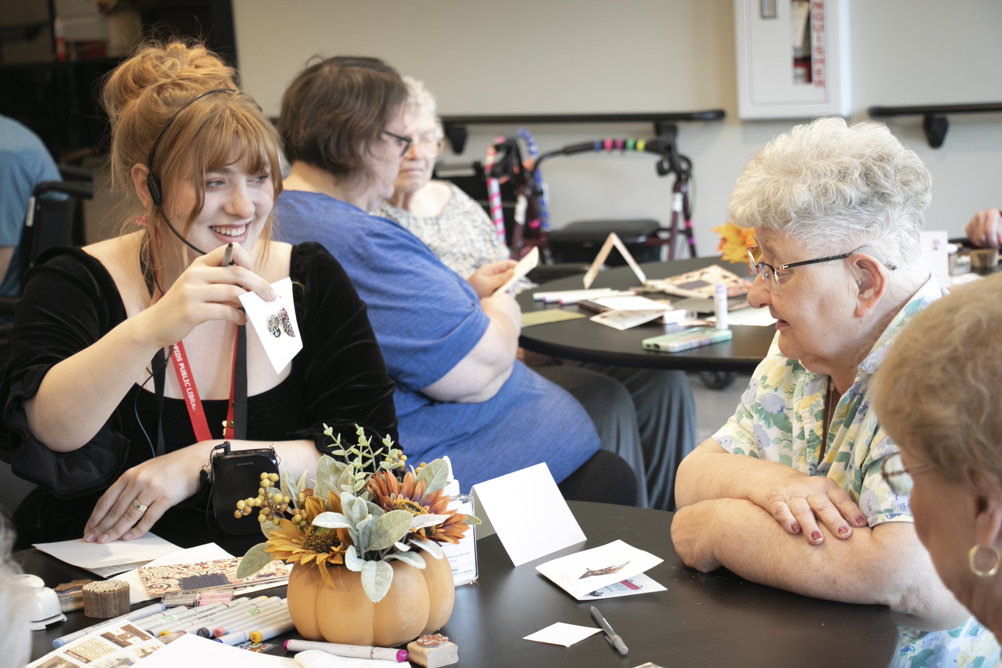 A woman, Erin, holds up a card with a stamp on it and smiles at two older women.