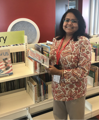 A woman in a red shirt holds a book in front of shelves.