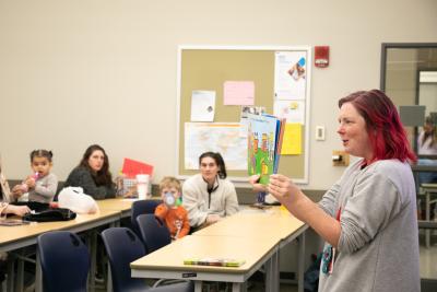 A woman holds up a children's book she is reading to a room of parents and children.