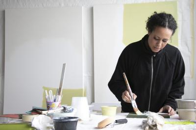 A woman, Akwi Nji, paints in front of partially finished canvases on a wall.