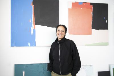 A woman, Akwi Nji, stands in front of her abstract paintings featuring blocks of color.