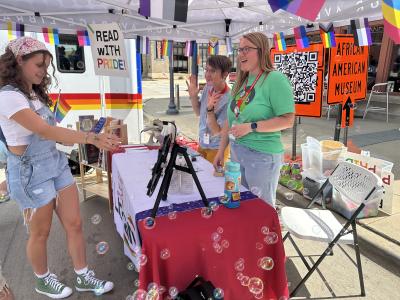 Two women smile behind a table and a sign that says "Read With Pride" as a third woman approaches.