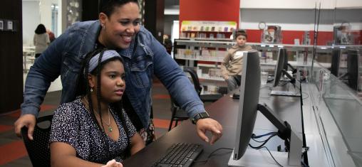 A woman smiles and points at a computer as a girl clicks with a mouse.