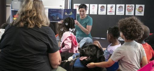 A woman reads a book to a room full of children and one dog, who wears a vest indicating he is a service dog.