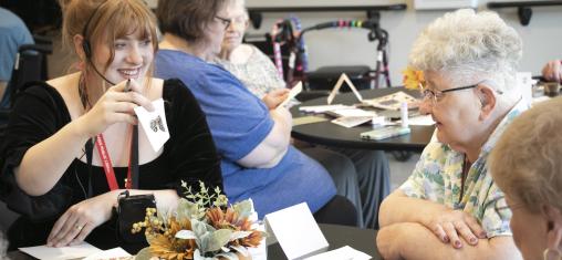 A woman, Erin, holds up a card with a stamp on it and smiles at two older women.