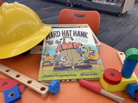 The book "Hard Hat Hank" sits on a table with a hard had and building toys.